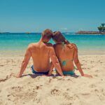 A couple enjoys a sunny day on the sandy seashore, surrounded by blue ocean and clear skies.